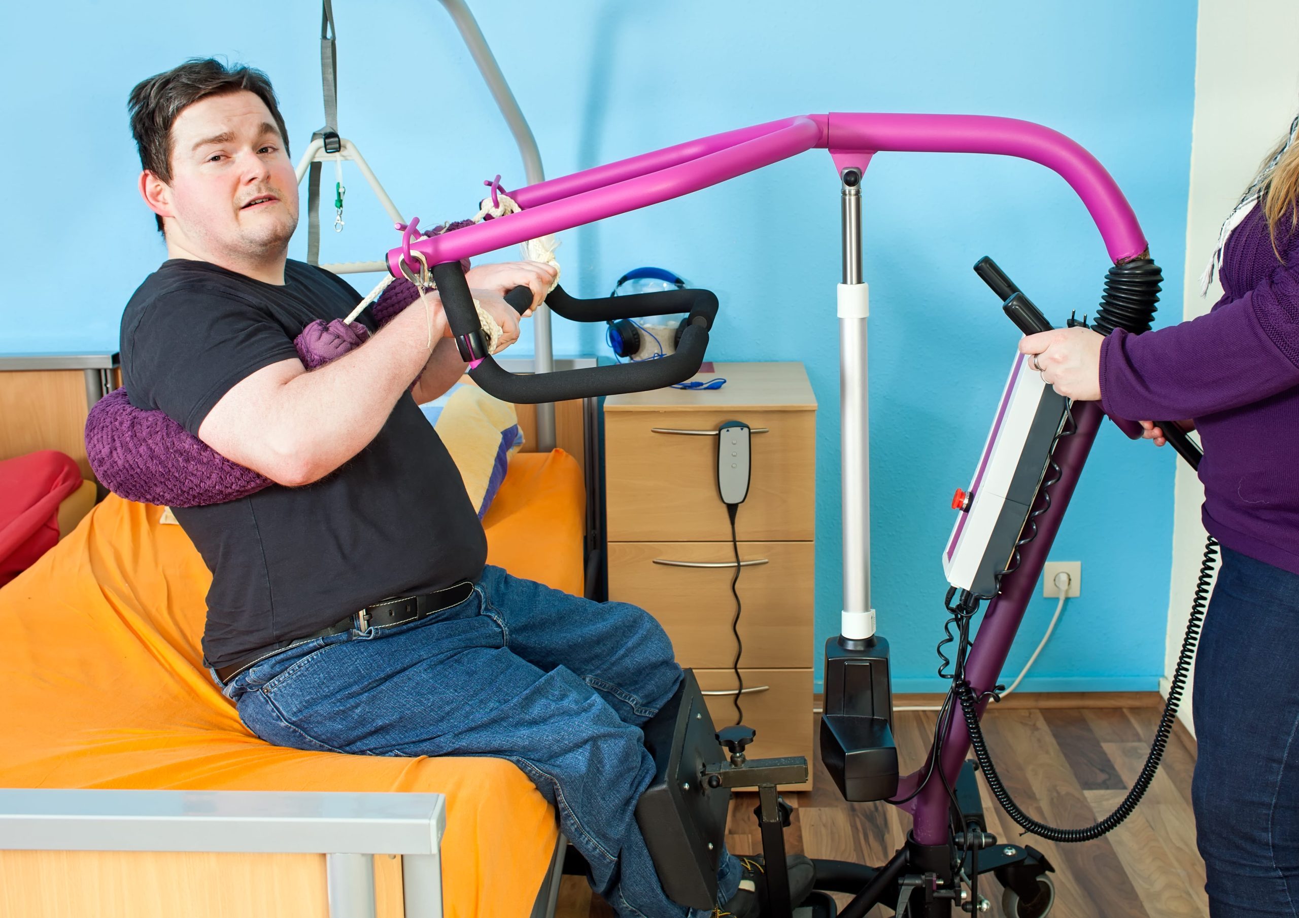 Spastic young man with infantile cerebral palsy caused by complications at birth using a patient lift to hoist himself out of a healthcare bed for transfer to a multifunctional wheelchair.