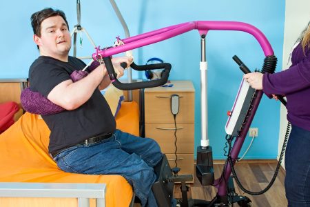 Spastic young man with infantile cerebral palsy caused by complications at birth using a patient lift to hoist himself out of a healthcare bed for transfer to a multifunctional wheelchair.